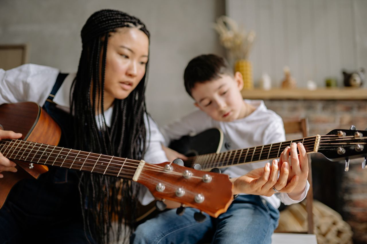 A mother helps her son learn to play guitar, fostering creativity and bonding.